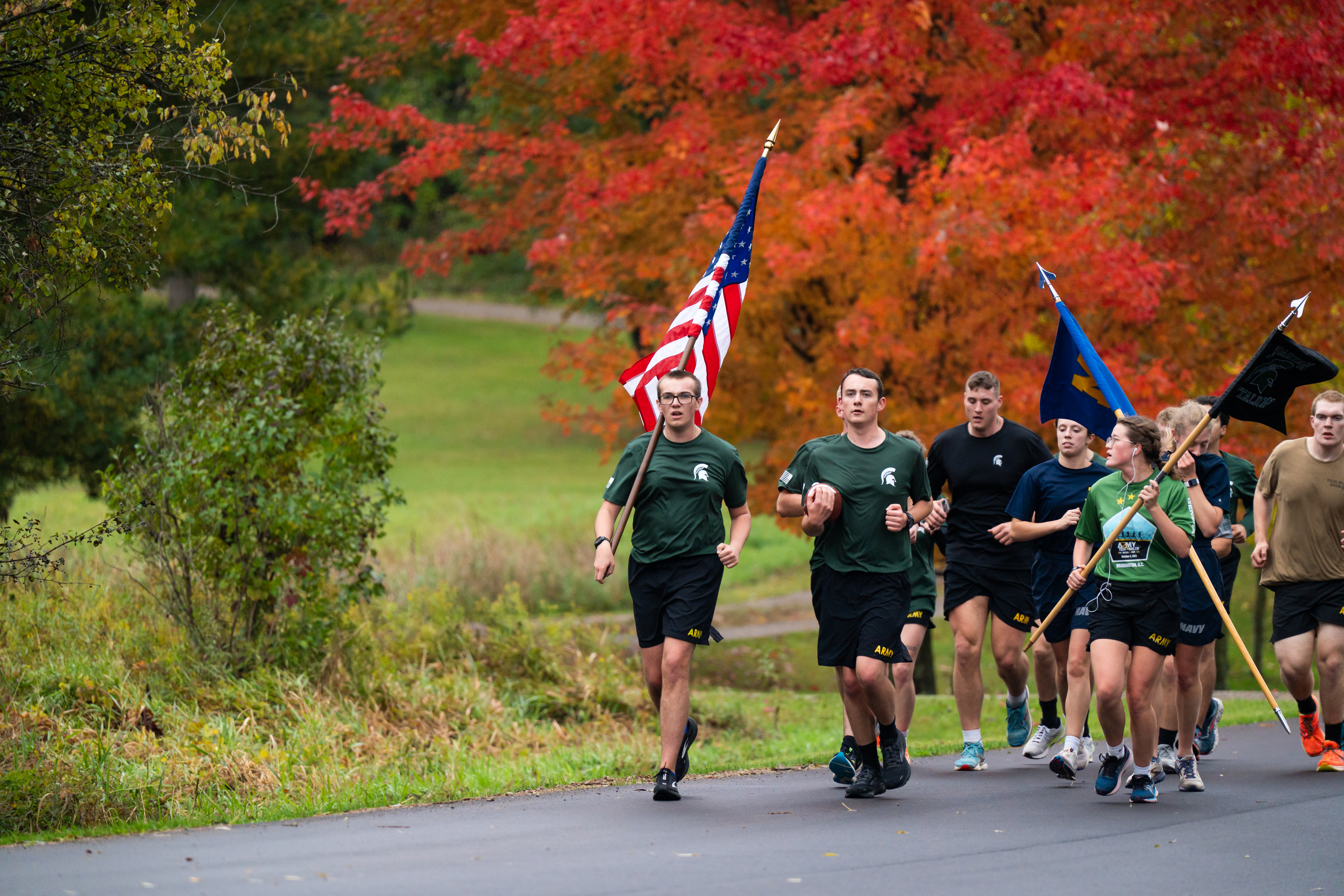 Army cadets running with US and Army flags with beautiful red/orange fall colors from the trees behind them