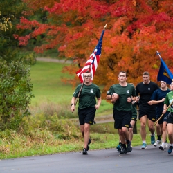 Army cadets running with US and Army flags with beautiful red/orange fall colors from the trees behind them